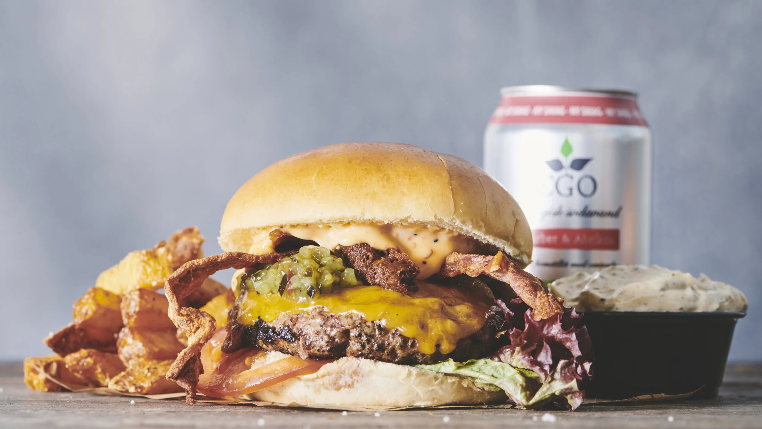 Couloir to-go En saftig burger med smeltet ost, bacon, salat og pickles står på et bord på Bistro Couloir. Ledsaget af sprøde chips, en lille skål dip og en drikkevaredåse giver baggrundens bløde grå nuance en rustik følelse til denne lækre take-away scene.