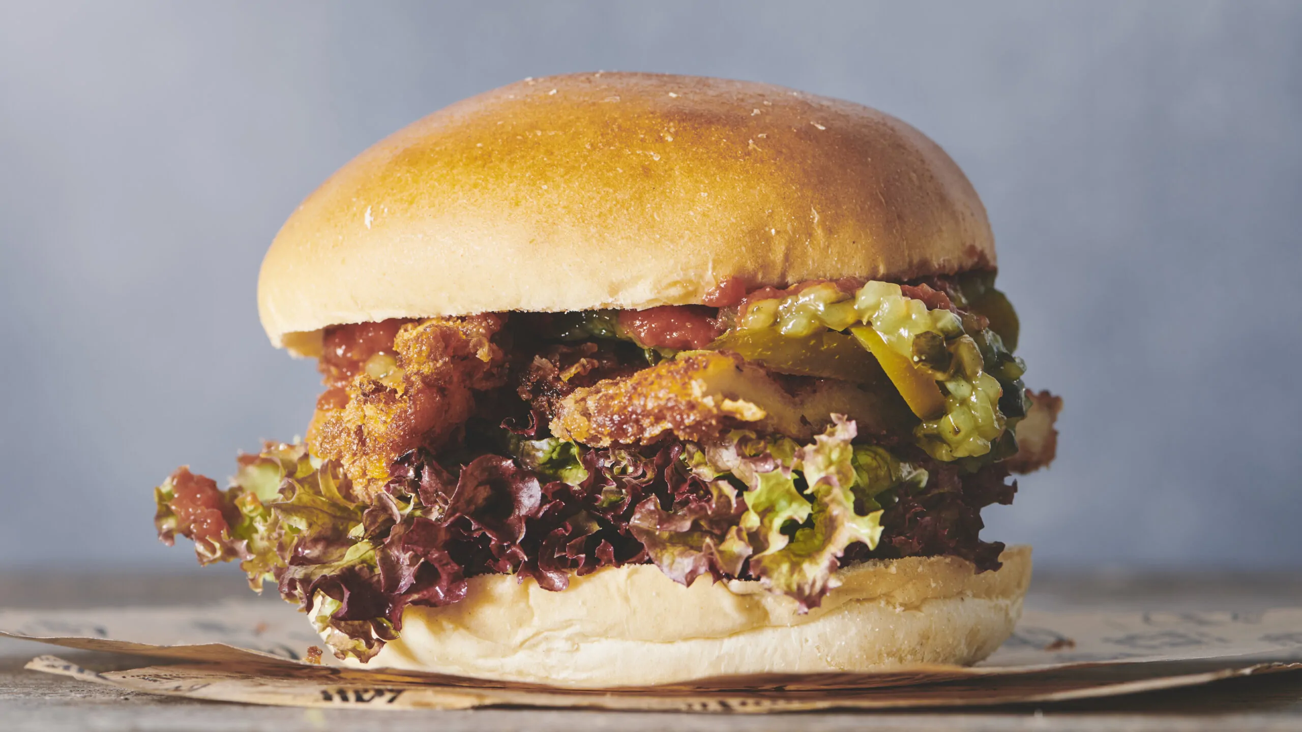 Couloir to-go Et nærbillede af en gourmetburger fra Restaurant Krimsvej med en gylden bolle, stegt kylling, salat, pickles og sauce hviler indbydende på en brun papiroverflade. Perfekt til take away eller nydes som dagens ret mod den slørede grå baggrund.